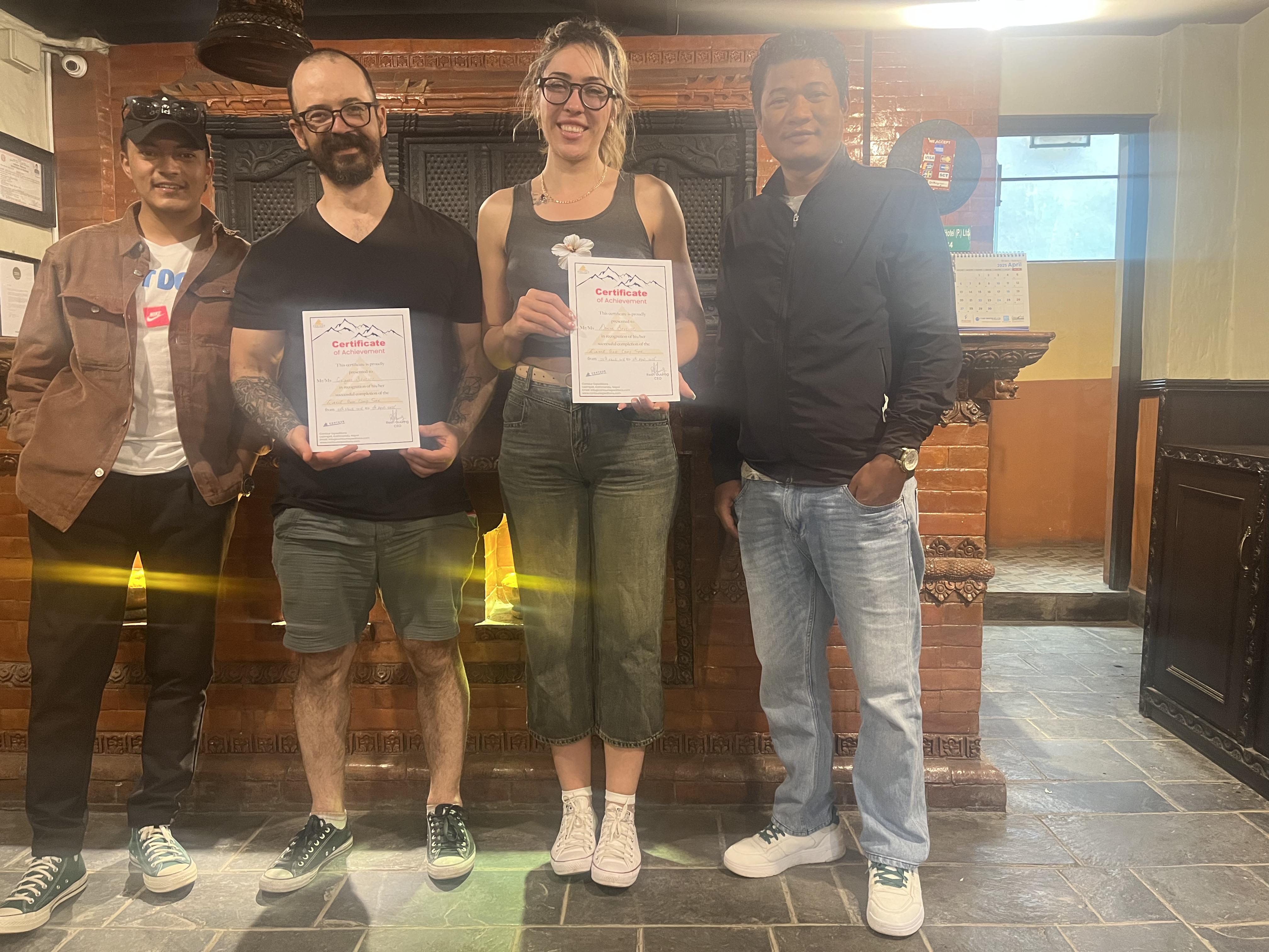 Four people posing indoors, with two holding certificates in front of carved wooden decor.